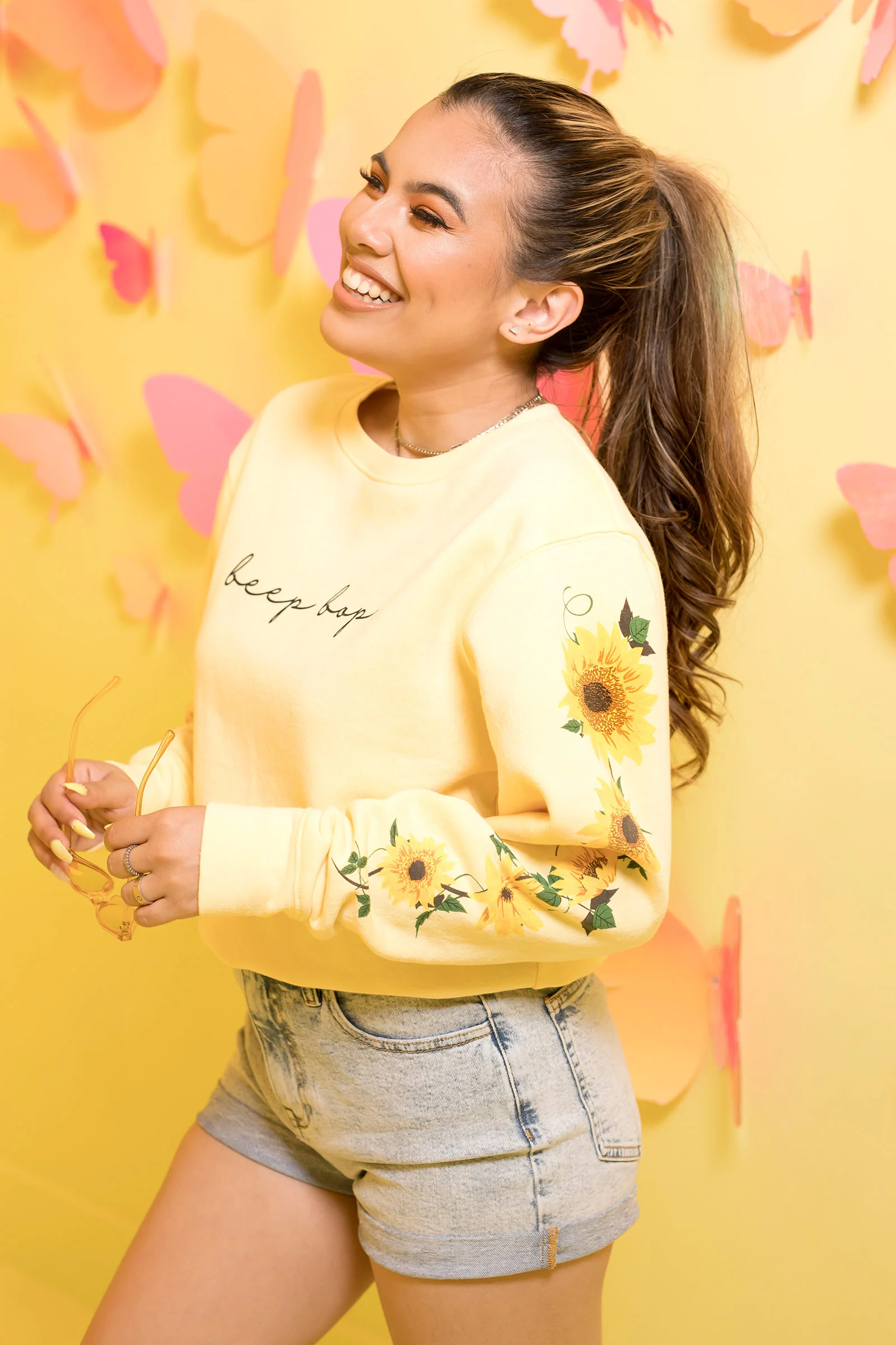 A smiling person in a yellow sweatshirt and denim shorts poses against a bright yellow wall with pink decorations.