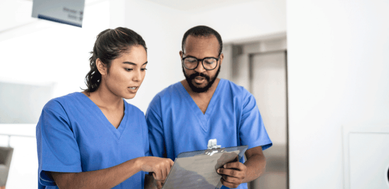 Two healthcare professionals in scrubs reviewing notes on a clipboard together.