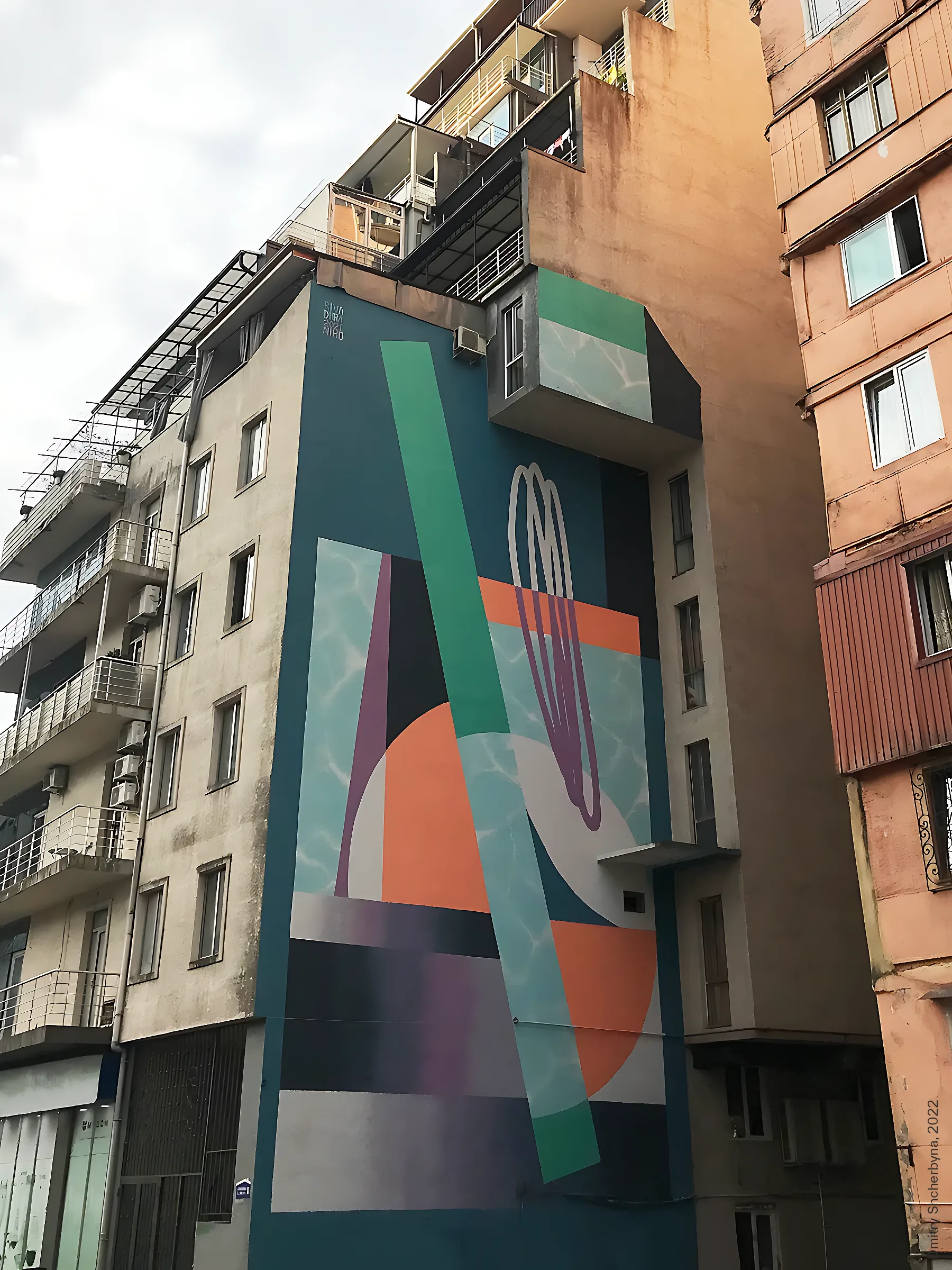 Abstract geometric mural painted in teal, orange, green and purple on the narrow side wall of a residential apartment block.