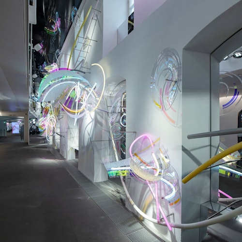 Instalação de arte moderna em ambiente interno, apresentando esculturas abstratas coloridas e iluminadas sobre paredes e pisos brancos em uma galeria espaçosa.