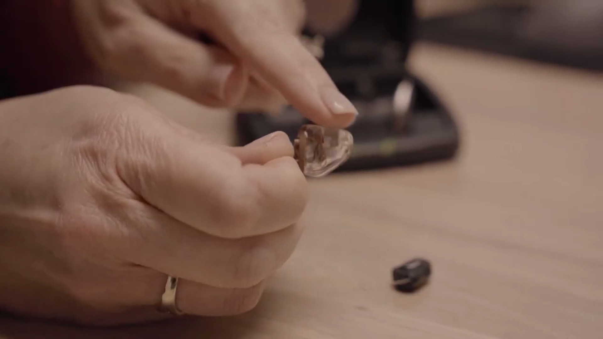 A close-up of hands working with small objects on a table, with a tool in the background.