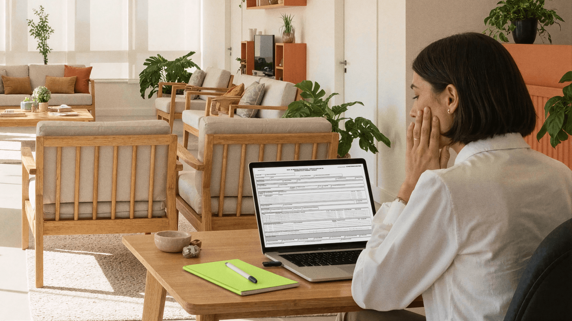 Profissional em área de recepção de clínicas analisa no notebook uma guia TISS, na Tivita; na mesa há um caderno verde e caneta, e ao fundo aparecem poltronas de madeira e plantas
