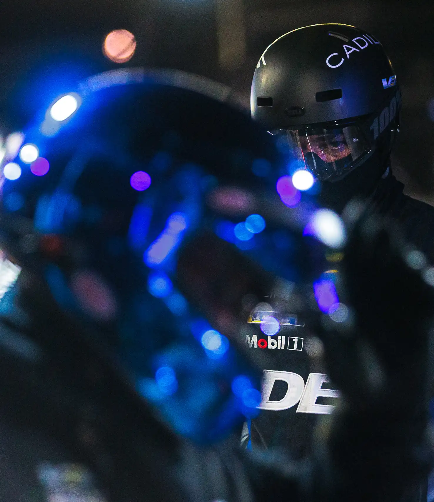 A Cadillac racing pit crew member in a helmet is sharply focused amid a blur of blue-lit reflections from another team member during a nighttime pit stop.