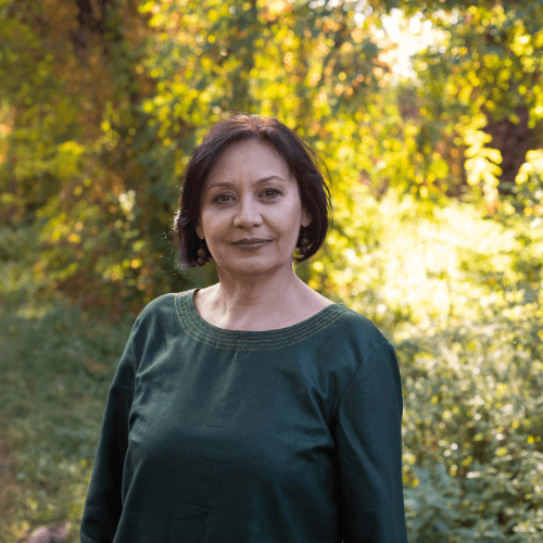 Portrait of a woman with pulled-back hair, wearing a textured sweater, seated in warm natural light against a soft brown background.