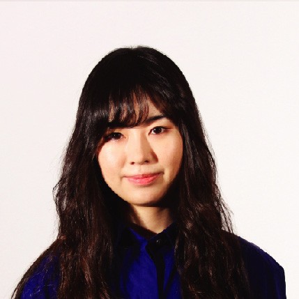 A young woman with long, dark brown hair and bangs wears a royal blue button-up shirt against a plain off-white background. Her gaze is directed at the camera, and she has a neutral expression.