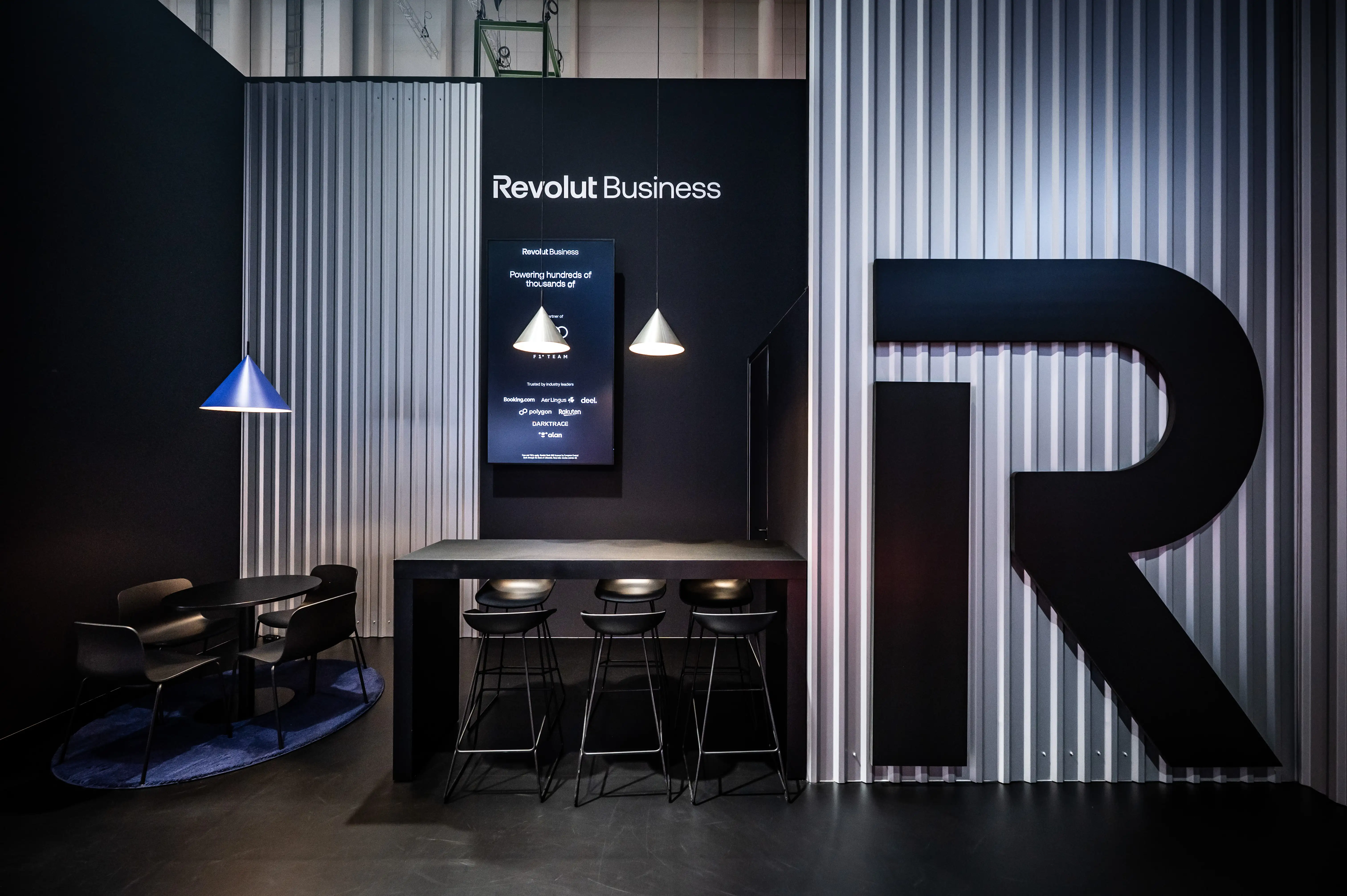 Revolut Business zone with black bar table, stools, screen and oversized R logo on corrugated metal wall.