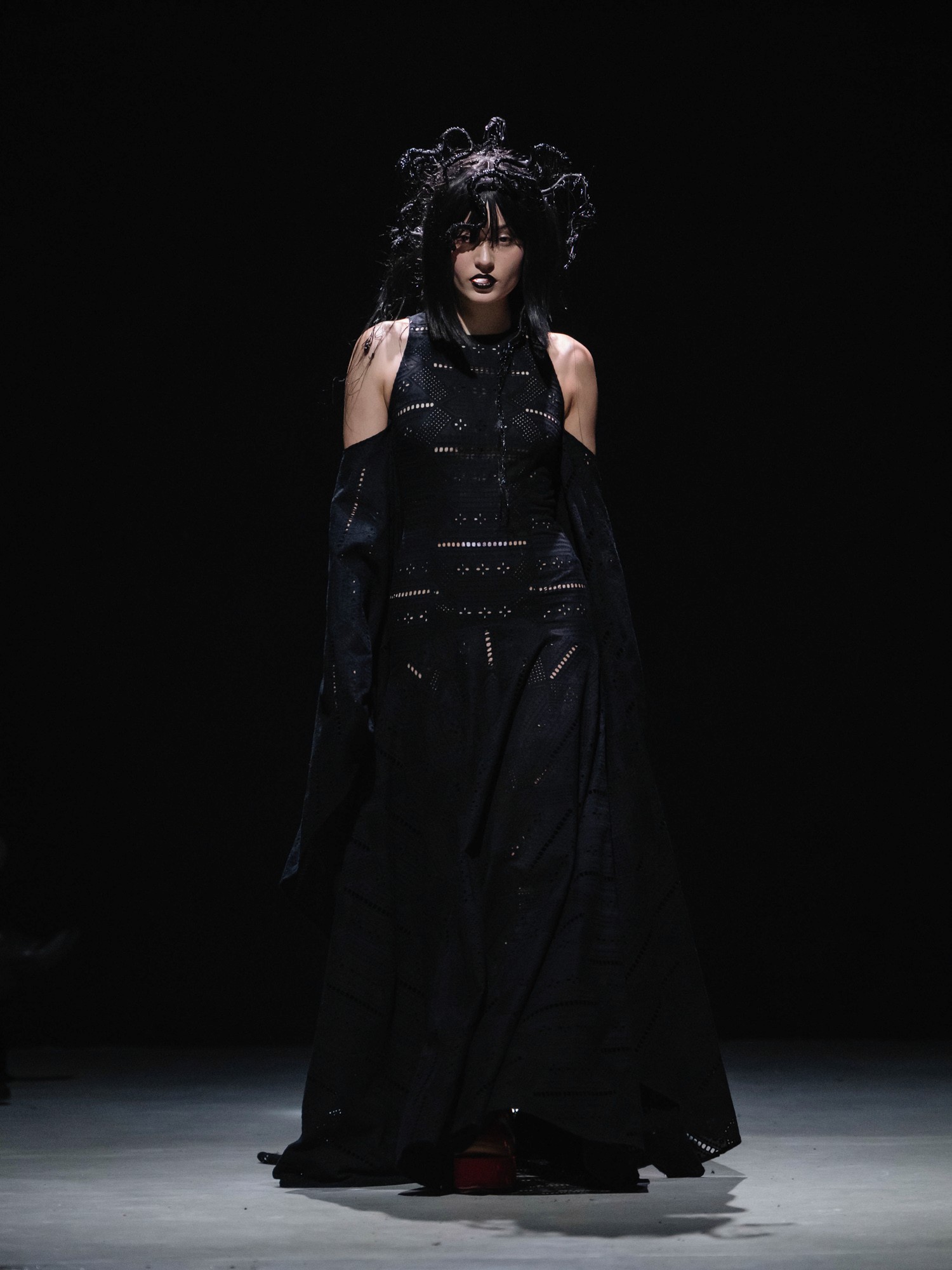A model walks the runway wearing a dramatic, gothic-inspired black gown with intricate cut-out patterns, complemented by striking dark makeup and a voluminous, intricate hairstyle, set against a dark, minimalist background.
