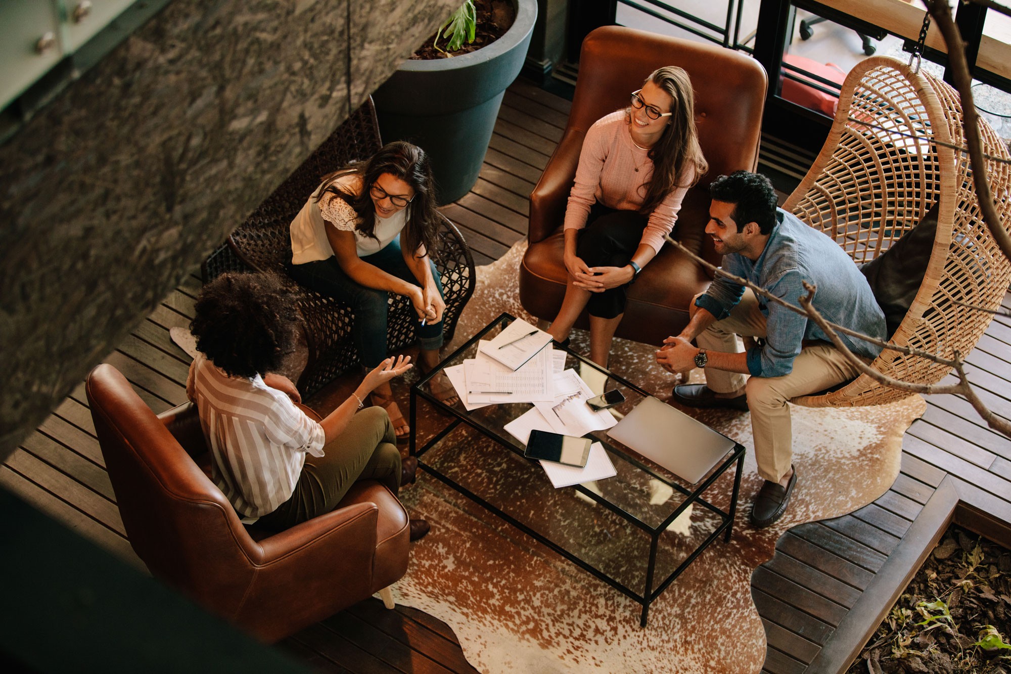 A group of four colleagues sitting in a cozy lounge area with comfortable chairs and a glass table, engaged in a casual discussion with documents and devices spread out on the table.