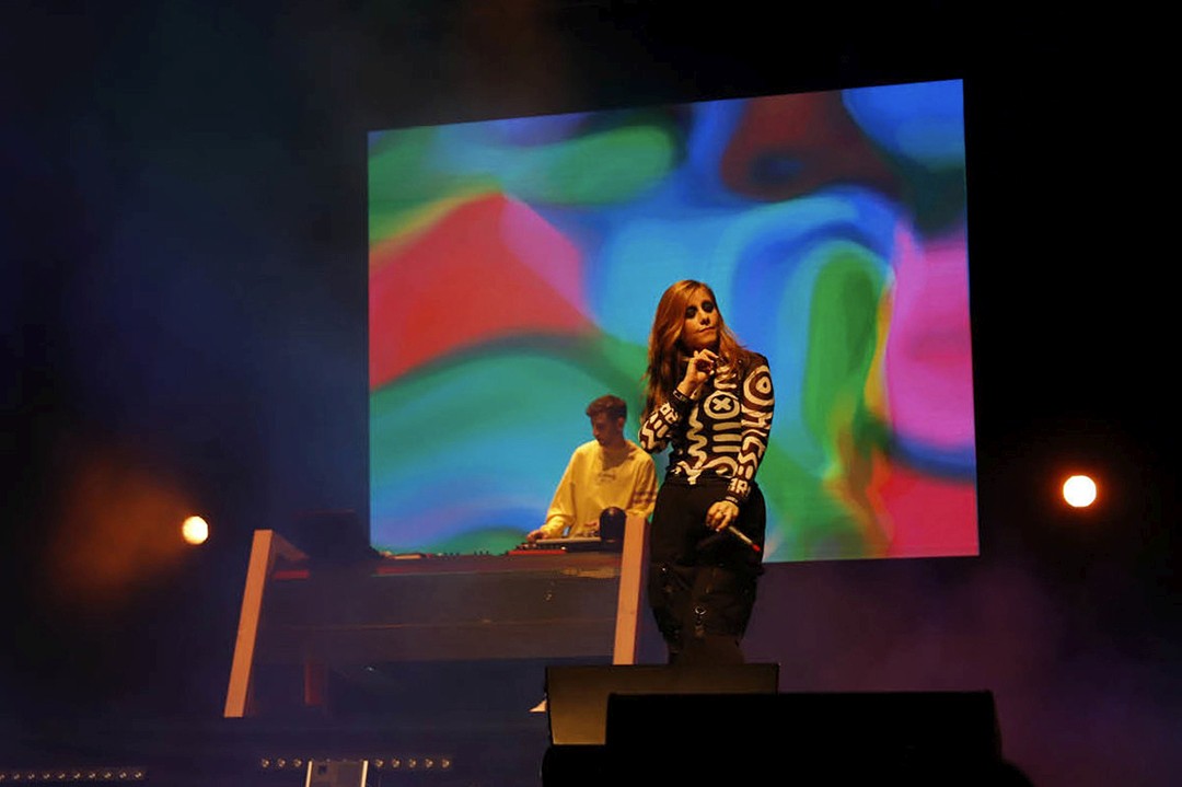 Laura Lamontage and Pico Amperio on stage. Large-format screen with fluid digital texture in pastel colours