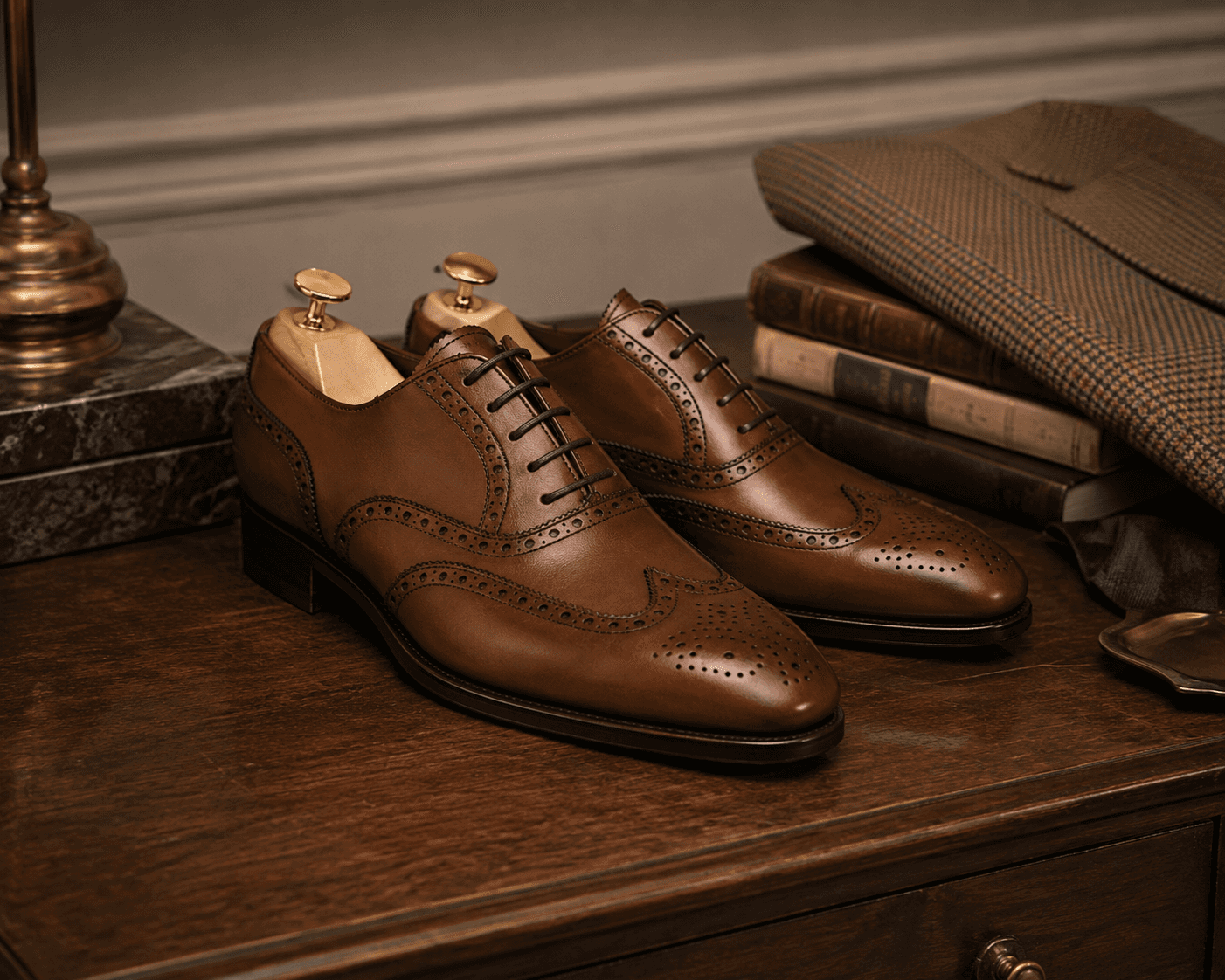 A pair of finely crafted chestnut brown leather full-brogue wingtip oxford shoes fitted with wooden shoe trees on a dark polished wooden table. The classic menswear arrangement features stacked vintage hardcover books, a folded houndstooth tweed jacket, a valet tray, and a brass table lamp under warm, atmospheric interior lighting.