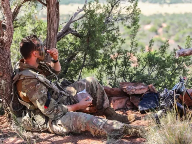 Soldier drinking from his canteen under a tree in a patrol base, ruck and gear beside him