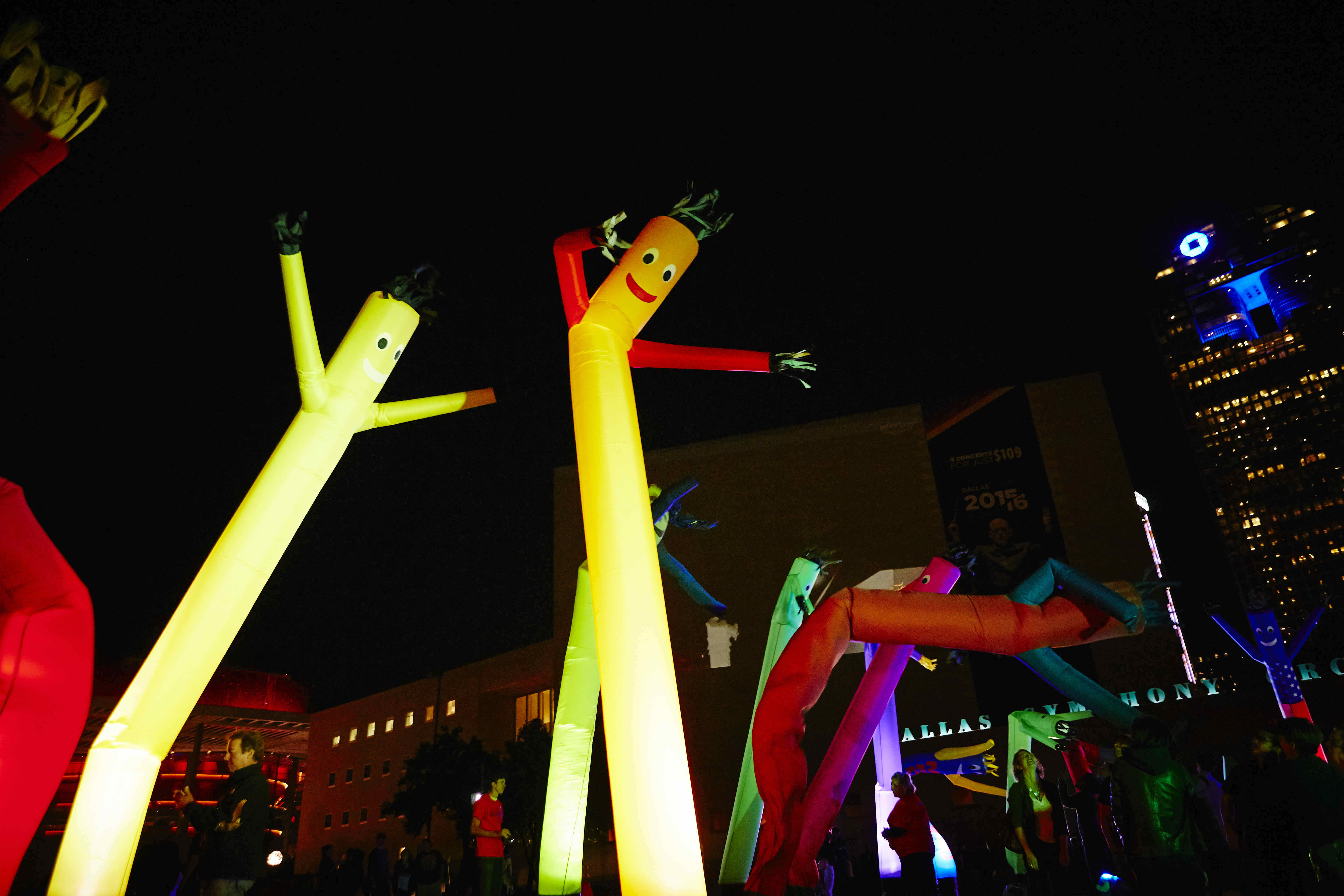 Brightly colored inflatable tube men dancing in a nighttime setting with colorful lights in the background.