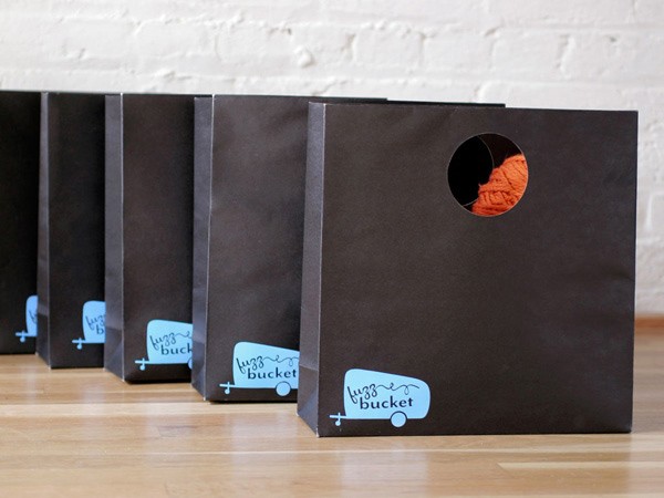 Four dark brown paper bags are lined up on a wooden surface. Each bag has a light blue chalkboard label that reads “fuzz bucket” with a small white drawing of a bucket at the bottom. A circular hole in the top right bag reveals an orange yarn or fabric inside. The bags are positioned against a white brick wall.