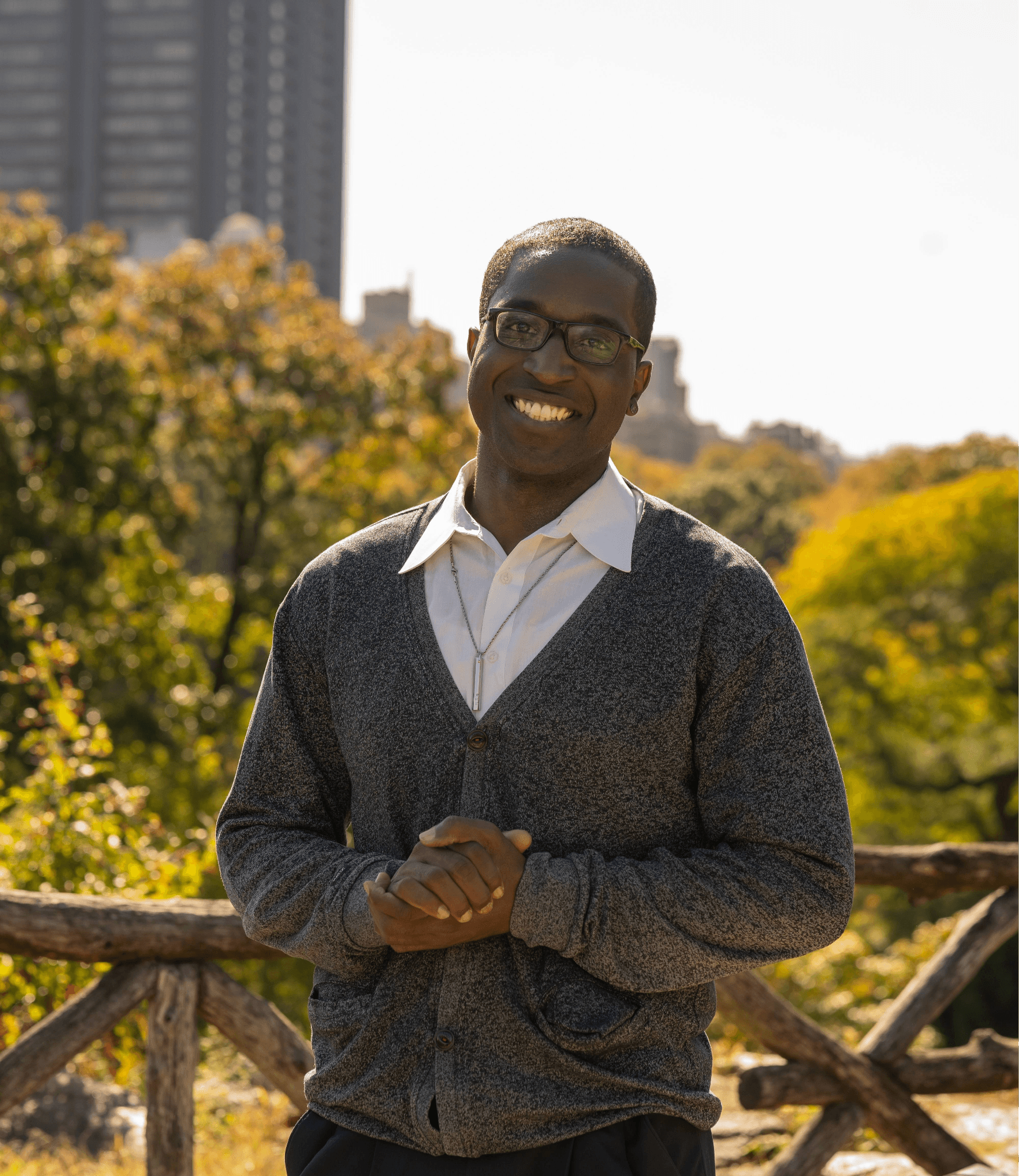 Jacob Laguerre smiling outdoors in a park, wearing glasses and a gray cardigan with autumn trees behind him.