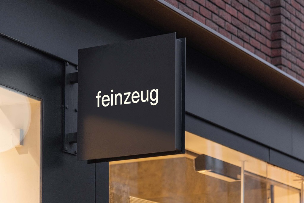 Modern architectural workspace with large floor-to-ceiling windows, surrounded by trees, and desks filled with design materials and books. Two blurred figures are walking through the space.