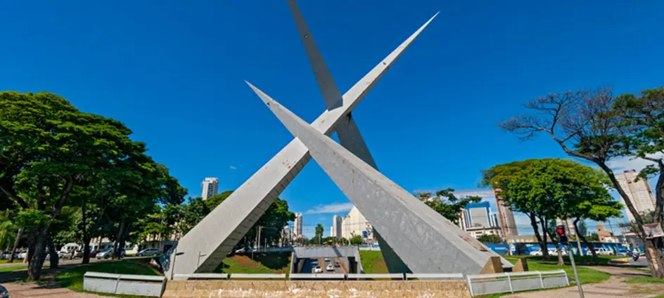 Fotografia panorâmica de Salvador, Bahia, em um dia ensolarado com céu azul e nuvens brancas. Em primeiro plano, à esquerda, aparece o Elevador Lacerda, estrutura alta e bege que liga a parte alta à parte baixa da cidade. Ao centro e à direita, vê-se a Baía de Todos-os-Santos com águas azul-turquesa, várias embarcações ancoradas e alguns navios ao fundo. Na parte baixa, destaca-se um prédio histórico amarelo com telhado de duas águas e, ao redor, outras construções e ruas movimentadas. Árvores verdes completam a paisagem urbana junto ao mar.