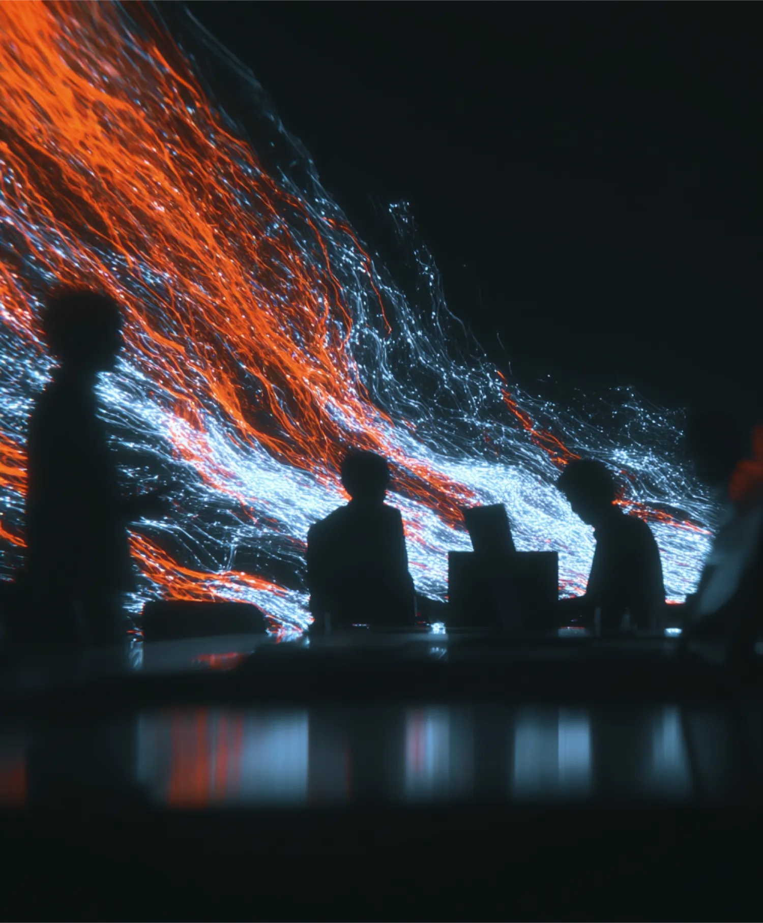 Silhouettes of people working at a table in front of a large screen displaying orange and blue abstract patterns.
