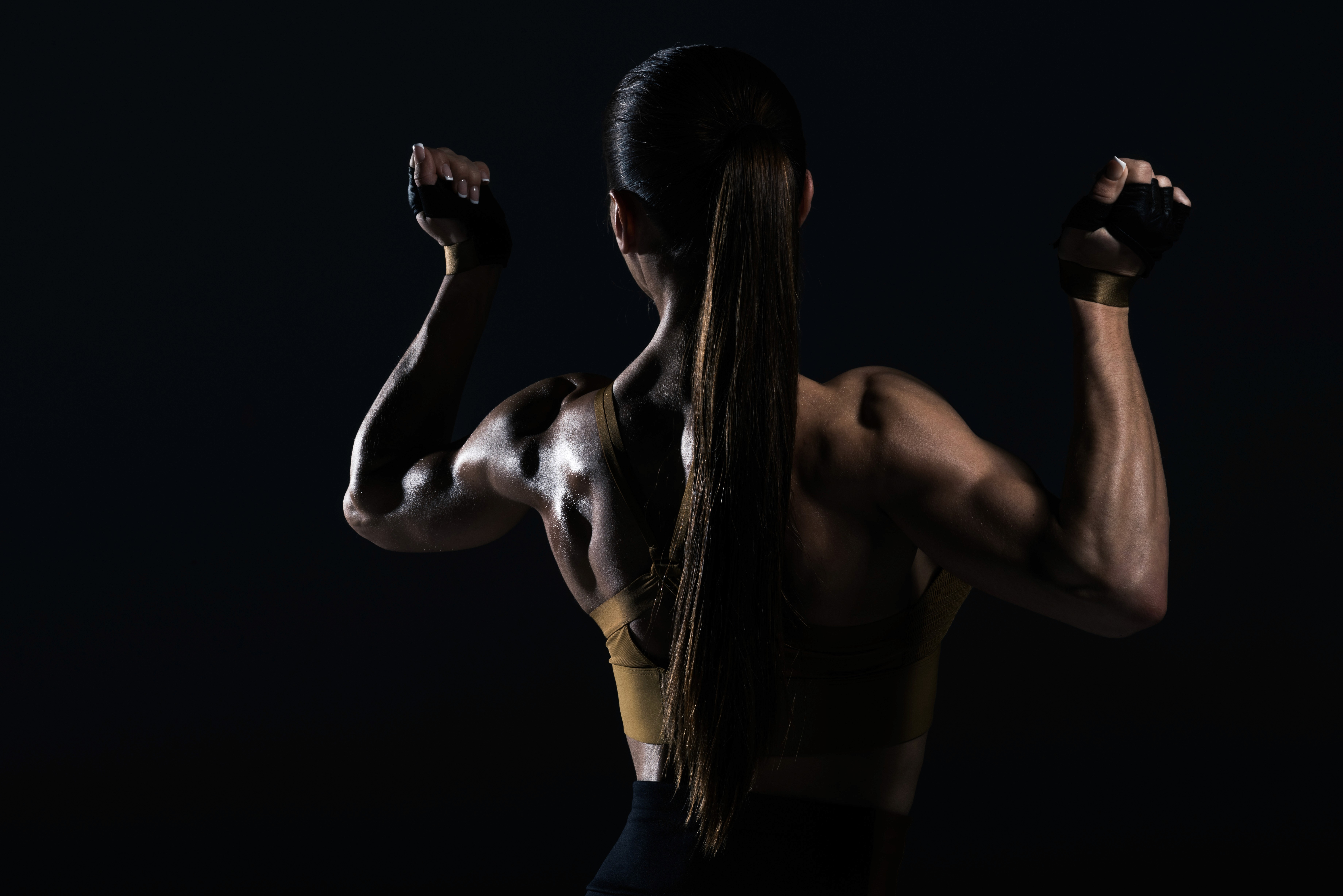 Fit woman with long hair and defined muscles flexing her arms, viewed from behind against a dark background, showcasing her strength and physique.