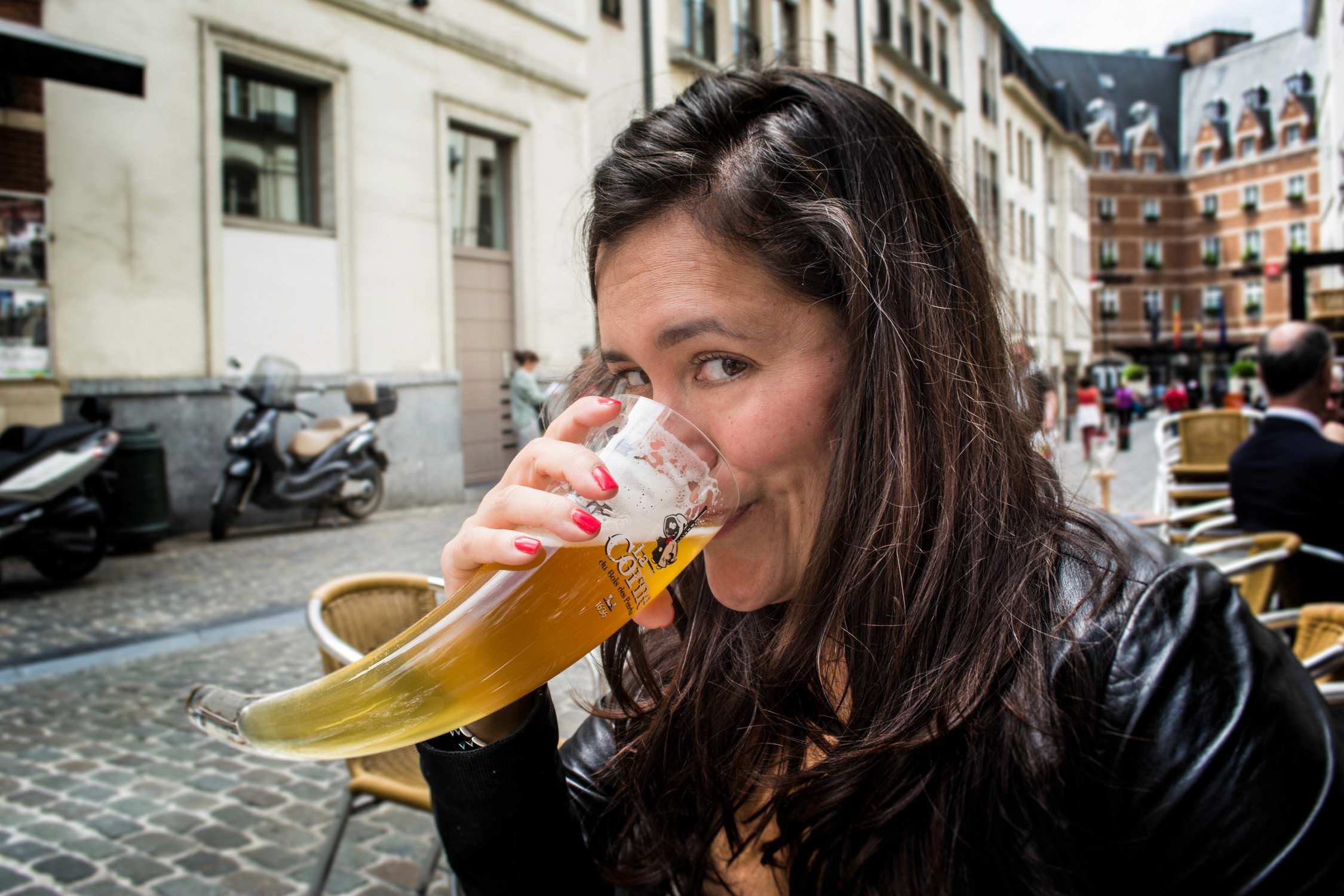 A woman with long dark hair smiles while holding a large glass of beer in front of a European street cafe.
