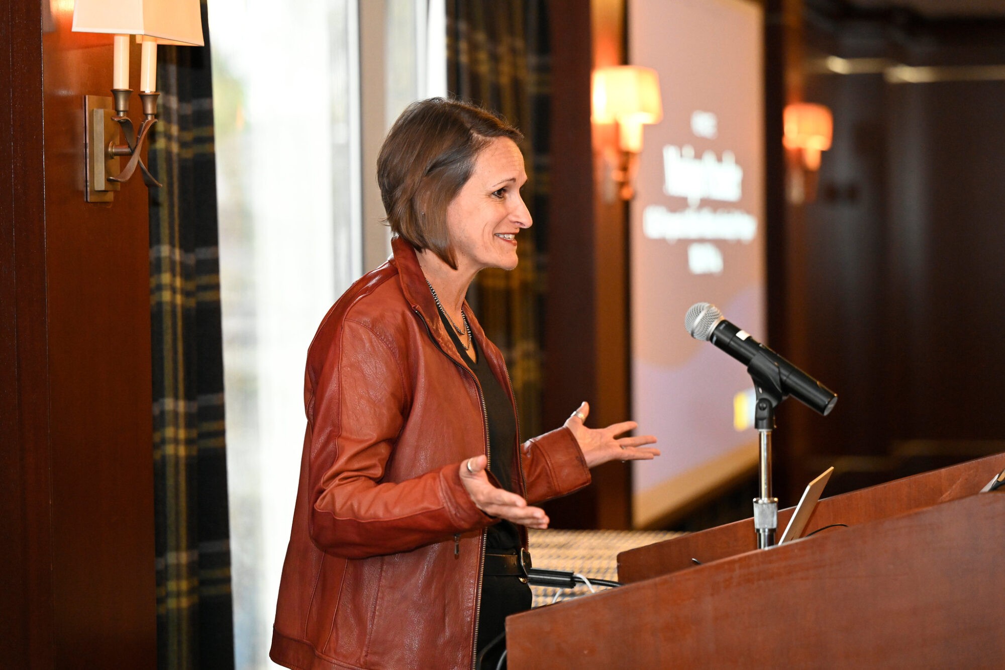 A woman gestures while speaking at a podium, with a microphone and warm lighting in the background.