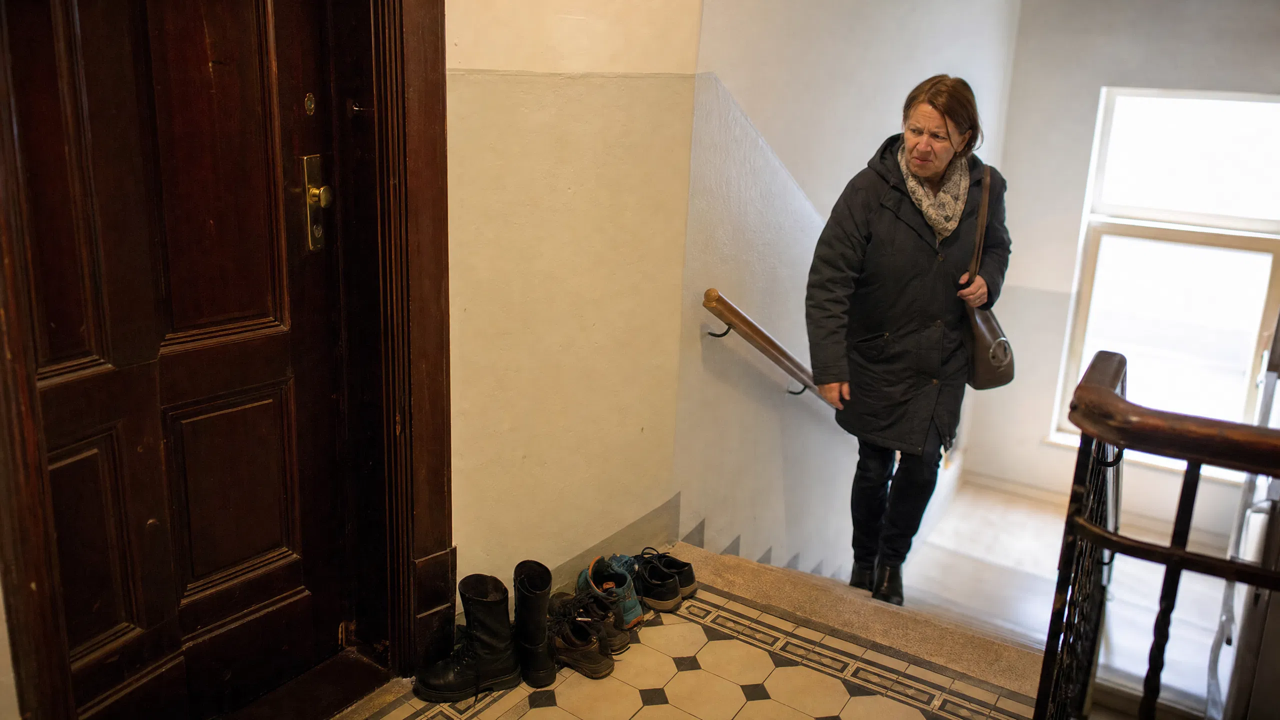 Eine Nachbarin geht im Treppenhaus eines österreichischen Altbaus die Stufen hinauf und blickt mit leicht missbilligender Miene auf mehrere unordentlich vor einer Wohnungstür abgestellte Schuhe und Stiefel.