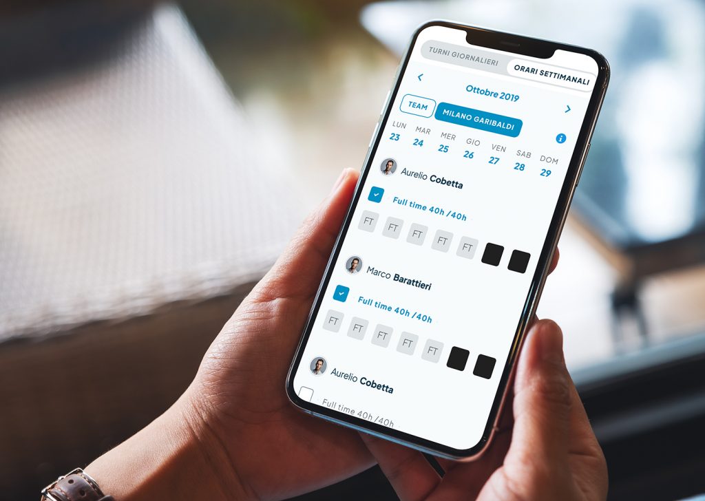 Una mano tiene uno smartphone aperto sulla schermata di gestione orari dipendenti.