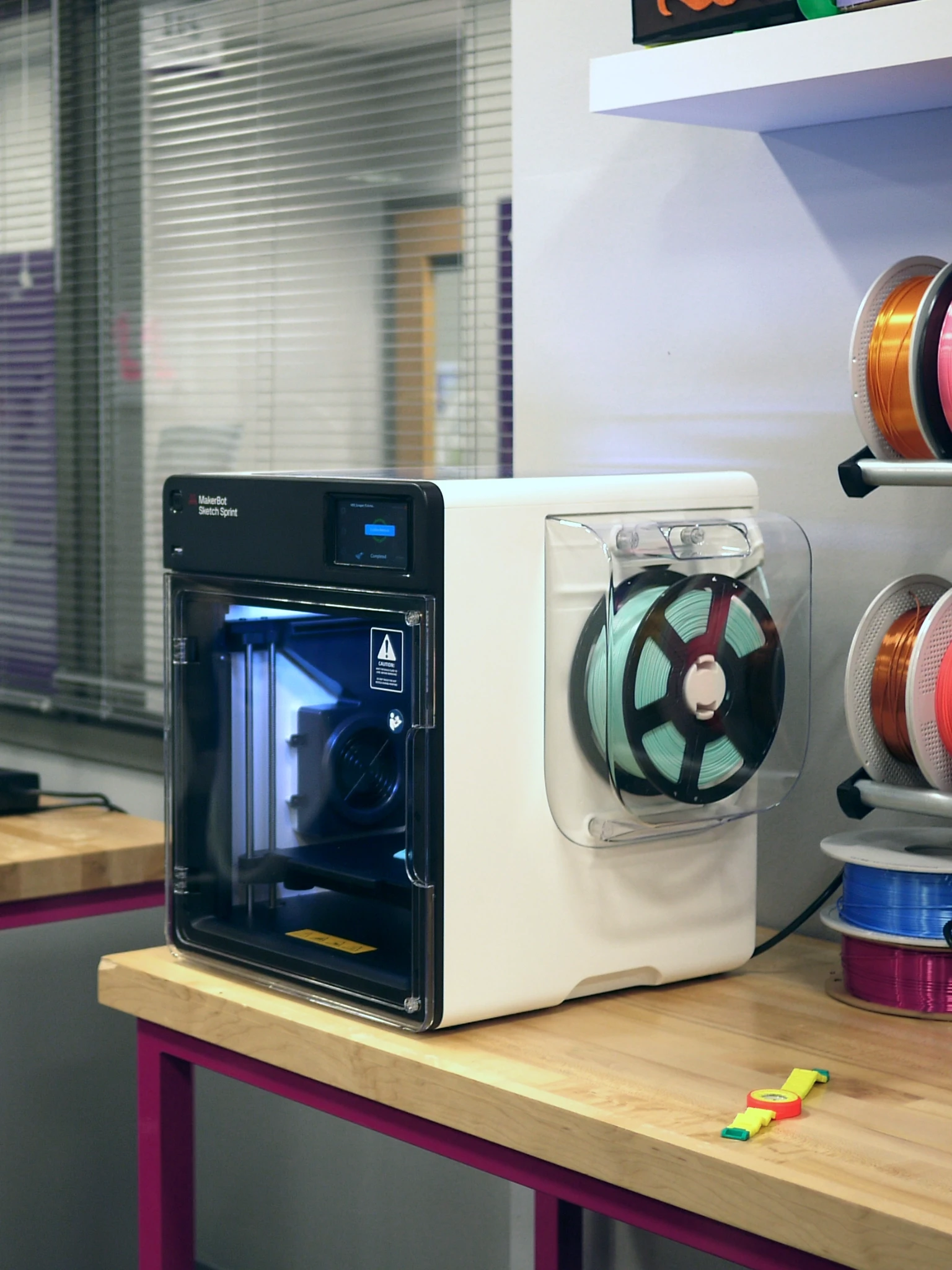 Modern home office setup with a monitor, ergonomic chair, keyboard, and indoor plants near a window with blinds.