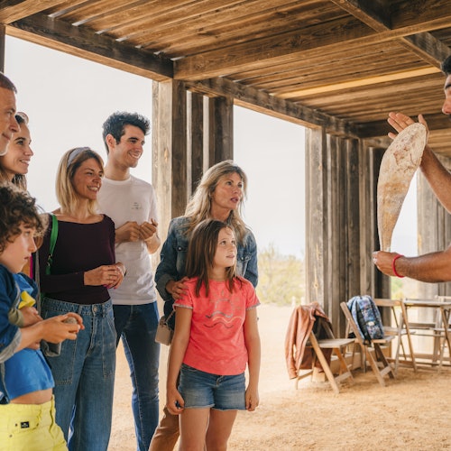 Eine Gruppe von Menschen beobachtet aufmerksam eine Person, die eine große Muschel unter einem hölzernen Pavillon hochhält.