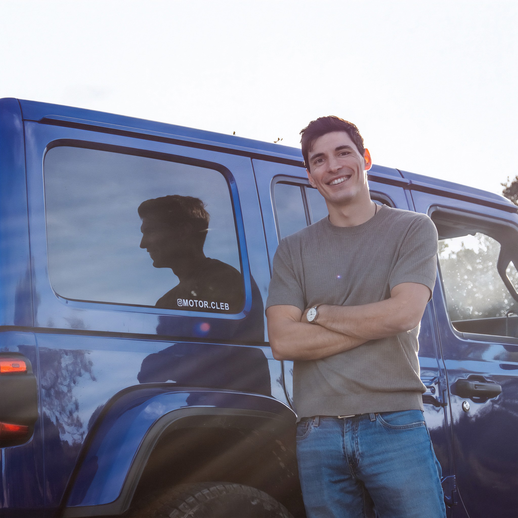 Two people in a workshop, one standing and smiling while the other leans over a car, working on it.