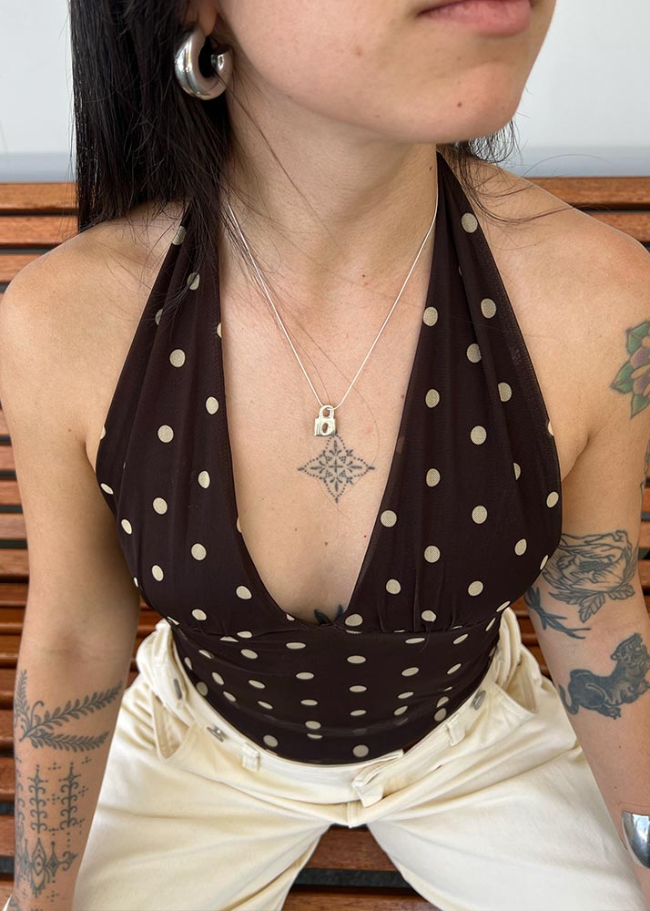 Person wearing a brown halter top with white polka dots, beige pants, and multiple tattoos. With a silver lock pendant.
