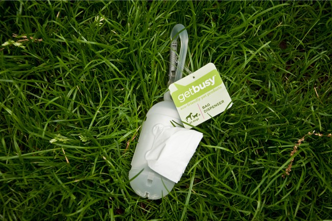 A white Getbusy bag dispenser lies on green grass. The dispenser has a green tag attached to a gray leash. The tag reads “getbusy” in white text, with “eco-friendly pet products” below it in smaller white text. A white tissue is partially visible inside the dispenser.