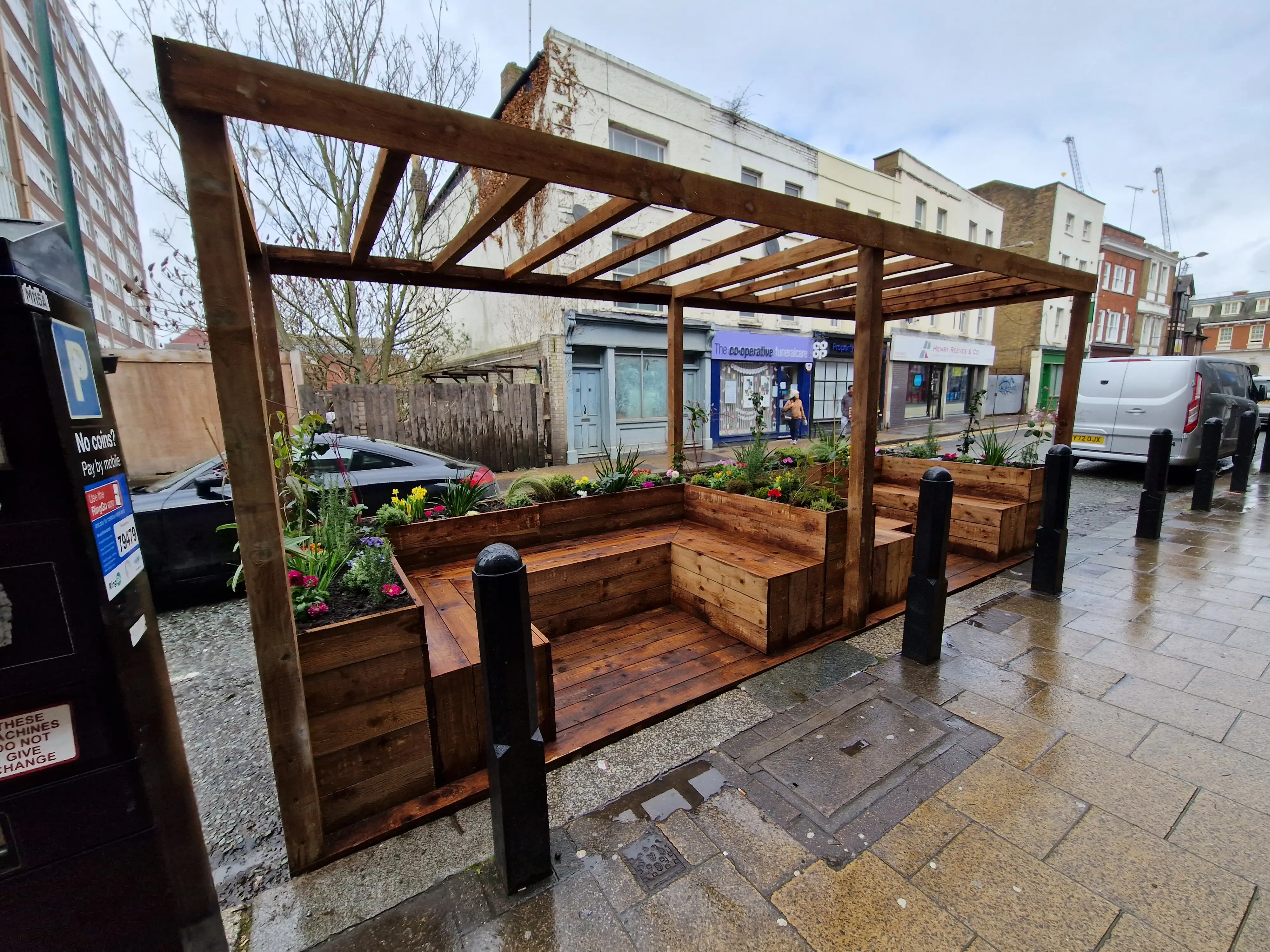 A wooden pergola structure with benches is set up in a public space, surrounded by pavement and buildings.