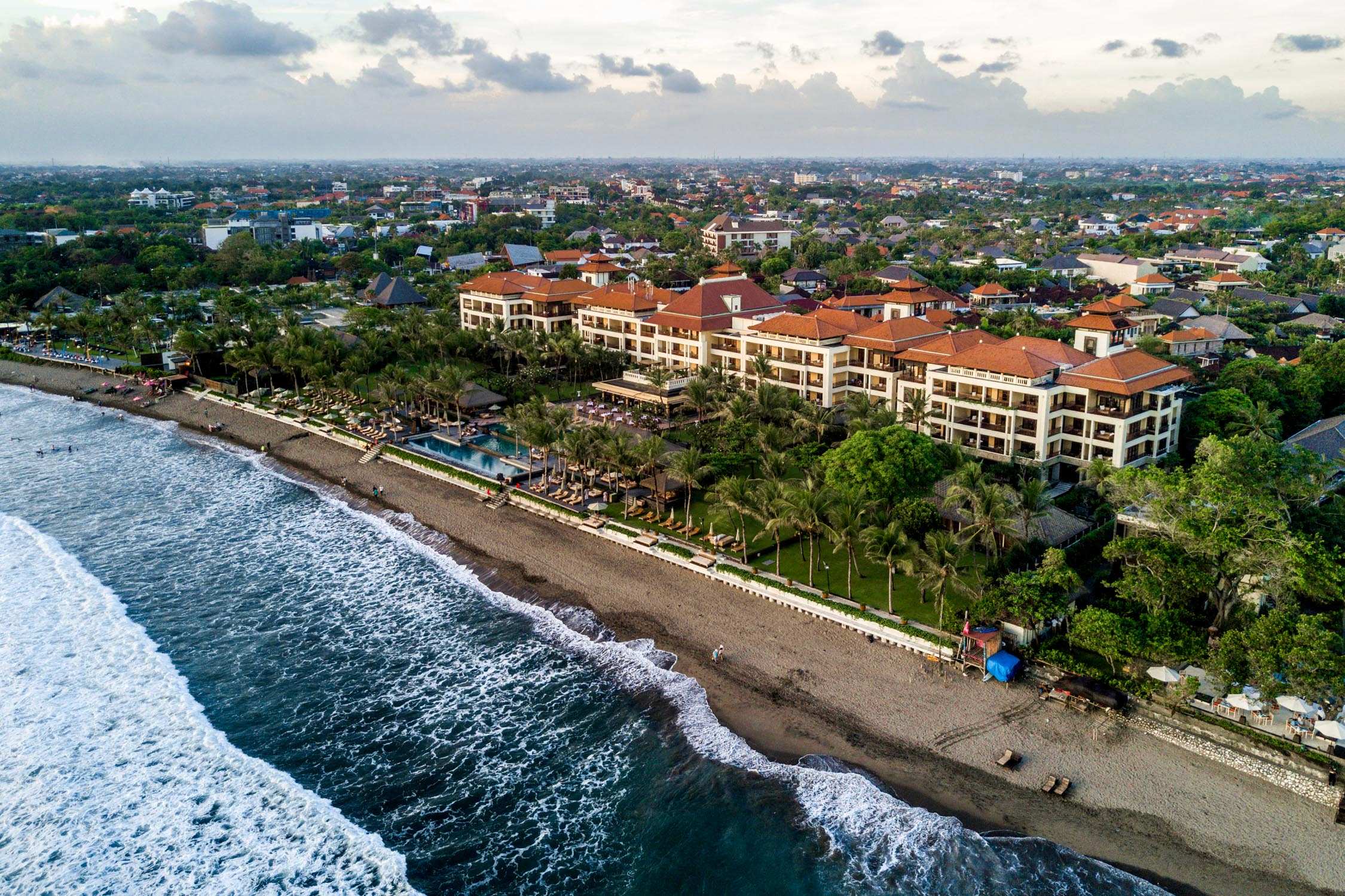 Aerial view of a Bali beach resort with terracotta roofs and swimming pools running down to the sand and turquoise water.