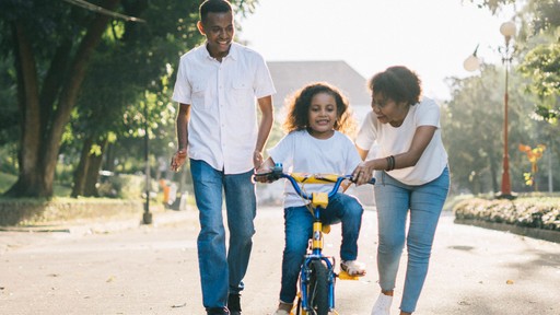 A family enjoys a sunny day outdoors, with a child riding a bike, assisted by two adults.