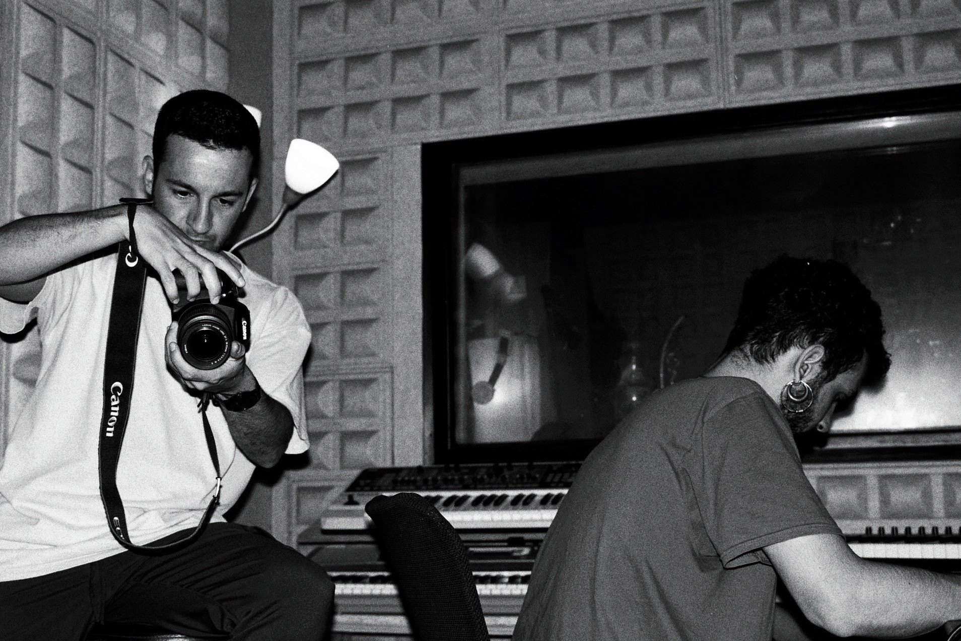 Two individuals working in a music studio, one on a computer and the other holding a camera, in black and white.