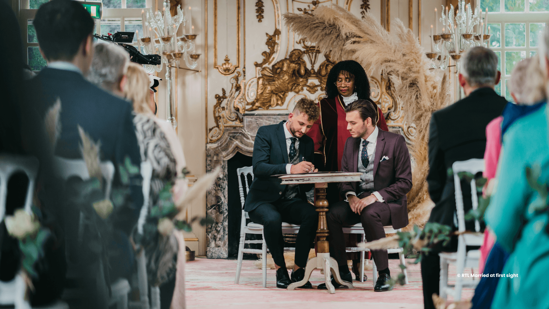 Een stel zit samen tijdens hun huwelijksceremonie, tegenover de trouwambtenaar en een elegante achtergrond. Gasten zijn aanwezig.