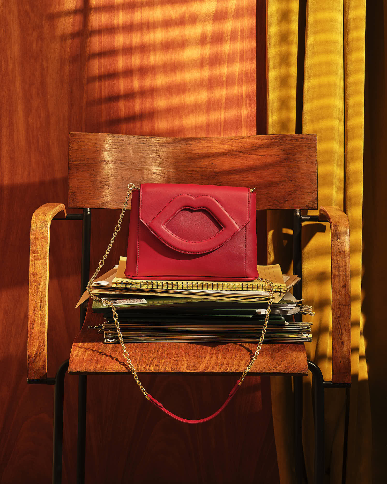 A red purse rests on a wooden chair in front of warm, striped lighting from blinds.