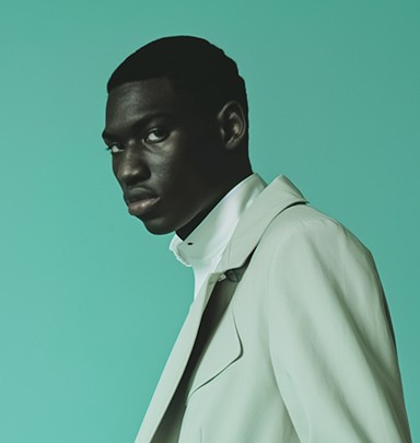 A man in a light-colored suit, gazing to the side against a green background.