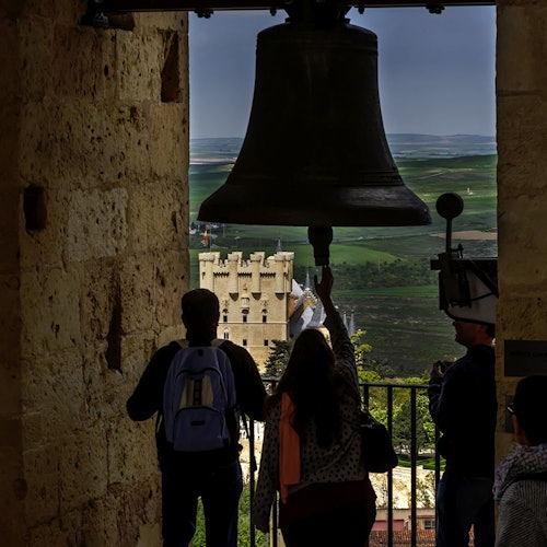 Three people stand under a large bell in a stone tower, looking out at a castle and green landscape in the distance.