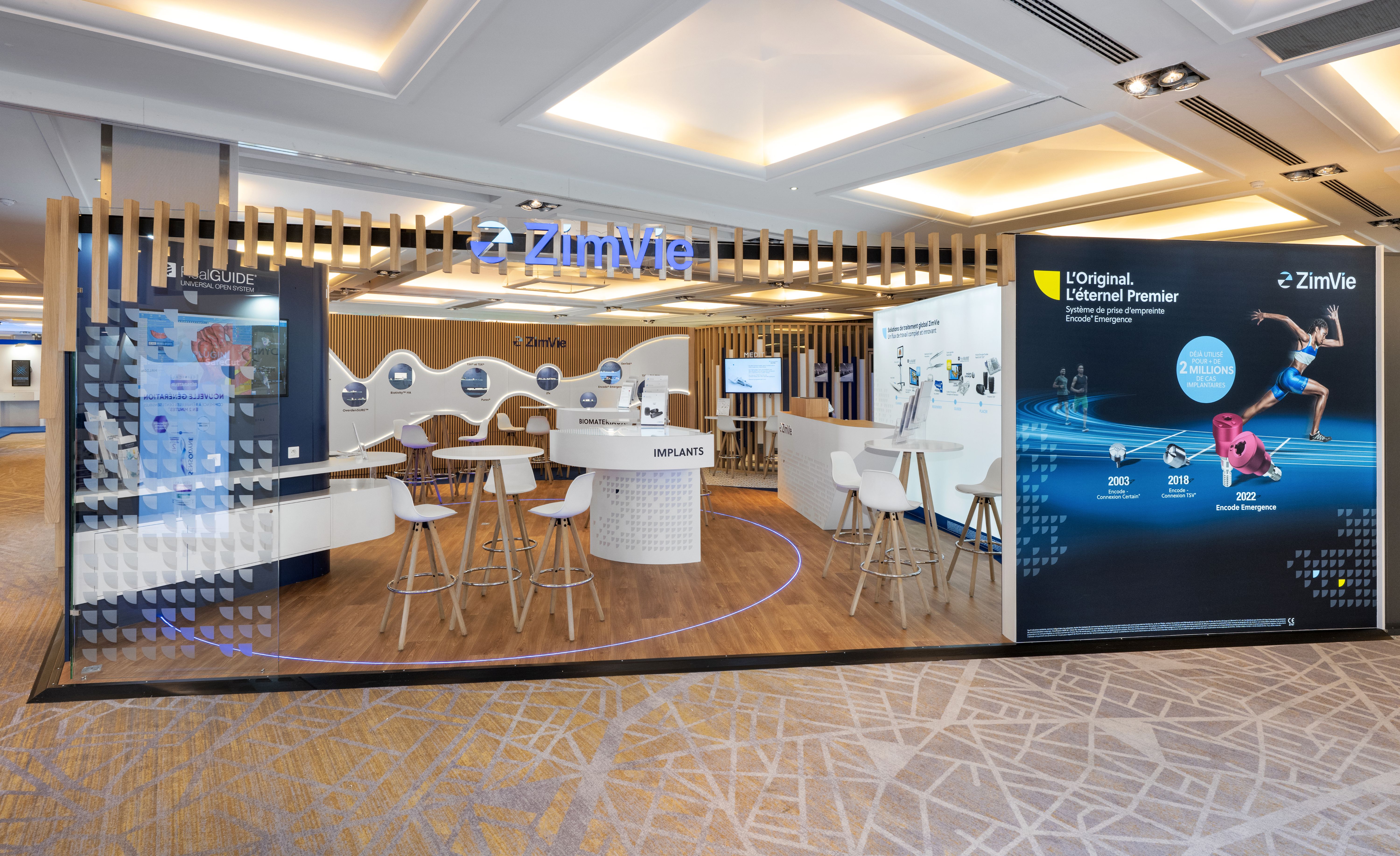 Stand ZimVie ADF 2025 conçu par Expace – vue extérieure avec logo lumineux bleu, comptoir Implants central, frise produits et panneau historique Encode Emergence L'Original L'Éternel Premier