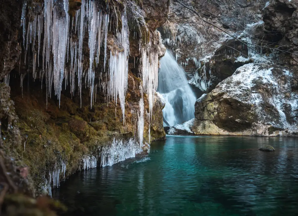  A frozen Šum Waterfall near the Vintgar Gorge in Slovenia, featuring sharp icicles hanging from a rocky moss-covered cliff over a deep emerald green pool during a cold winter day.