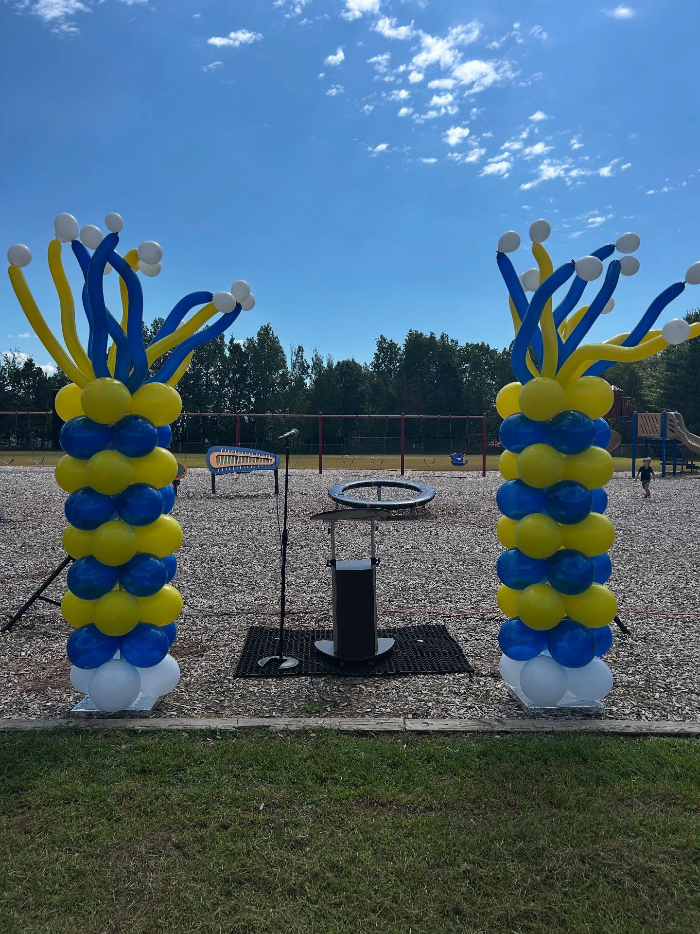 Outdoor setup with two large yellow and blue balloon columns featuring curly balloon accents designed by Be Bold Balloons