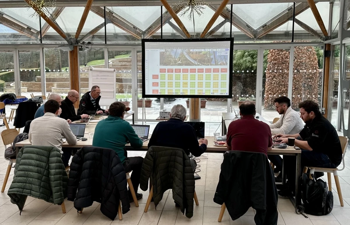 Group of people seated around a table with laptops during a workshop, facing a large screen displaying a grid of colored cells in a bright glass-walled room.