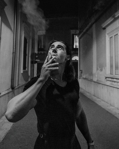 Man in black shirt standing in a narrow street at night, exhaling smoke in a cinematic black-and-white scene that conveys sophistication and depth.