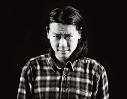 A black and white headshot of a man with long dark hair, wearing a plaid shirt, against a black background. His expression is serious, and his gaze is directed at the camera.