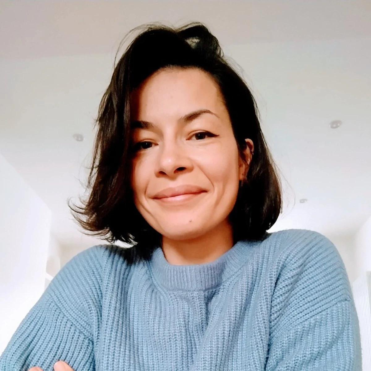 A woman with shoulder length dark brown hair smiles at the camera, she is wearing a light blue jumper and has her arms folded