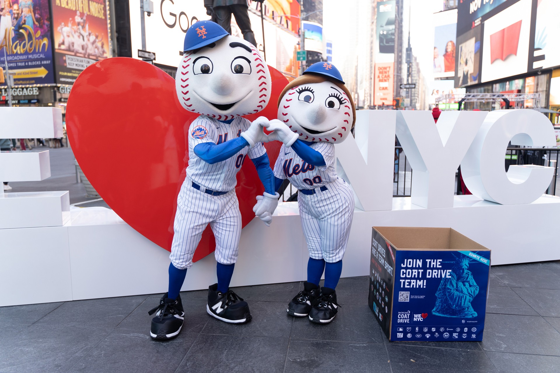 Mascots for the NY Mets baseball team post in front of the We Love NYC sculpture in Times Square. To their right is a donation box for the Coat Drive.