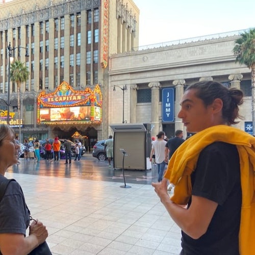 People walking near El Capitan Theatre with a brightly lit marquee at dusk, surrounded by tall buildings and palm trees.