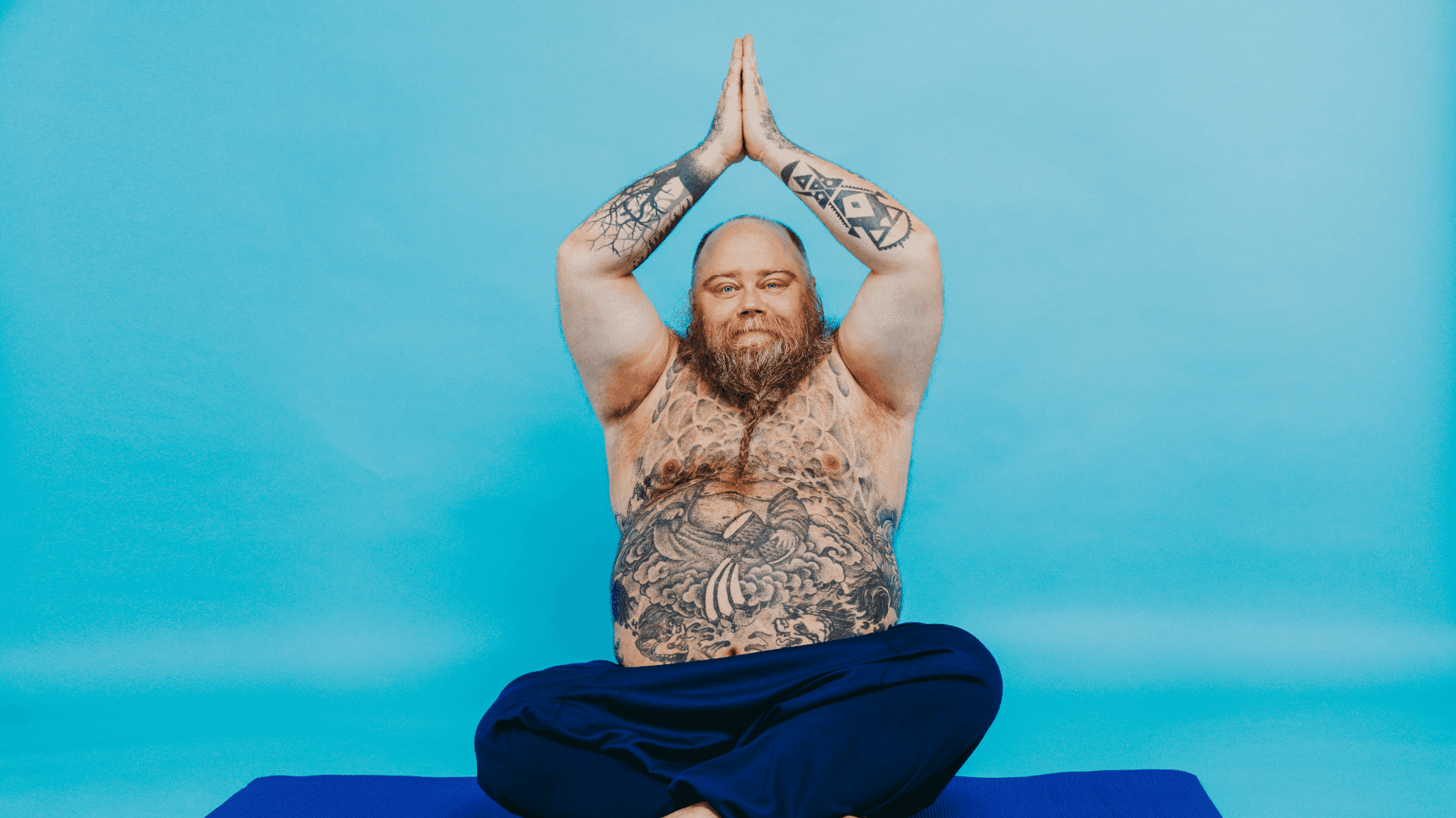 A tattooed person sits cross-legged on a yoga mat, hands raised in a prayer position against a blue background.