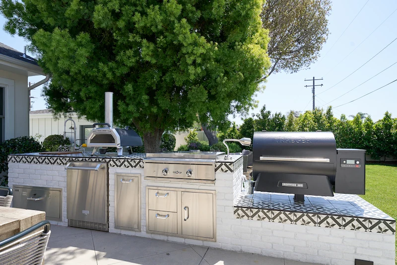 Outdoor BBQ station with patterned white tiles, stainless steel grill, under a large green tree. Costa Mesa Exterior Remodel by Todd Huge.
